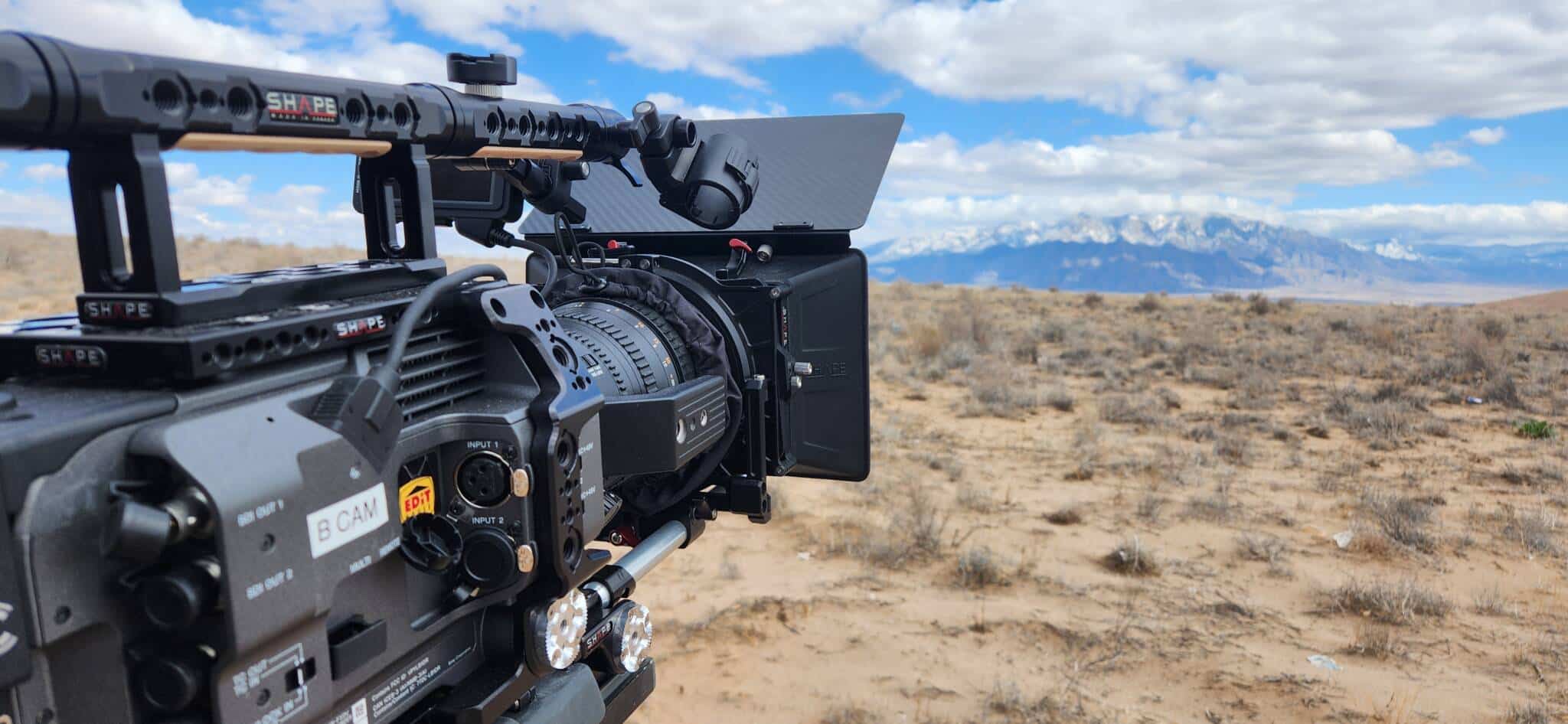 Close-up of a professional cinema camera on a tripod in a desert with mountains in the distance, blue sky above.