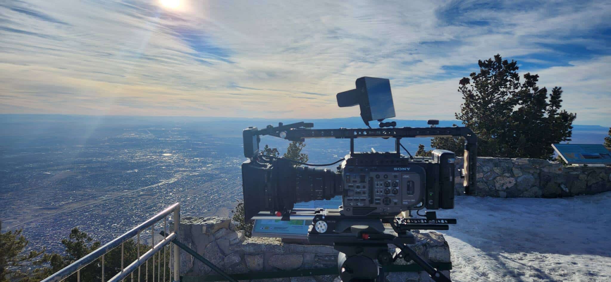 Professional cinema camera rig mounted on a tripod at a cliff overlook, aiming toward the expansive sea with a railing and stone wall in the foreground.