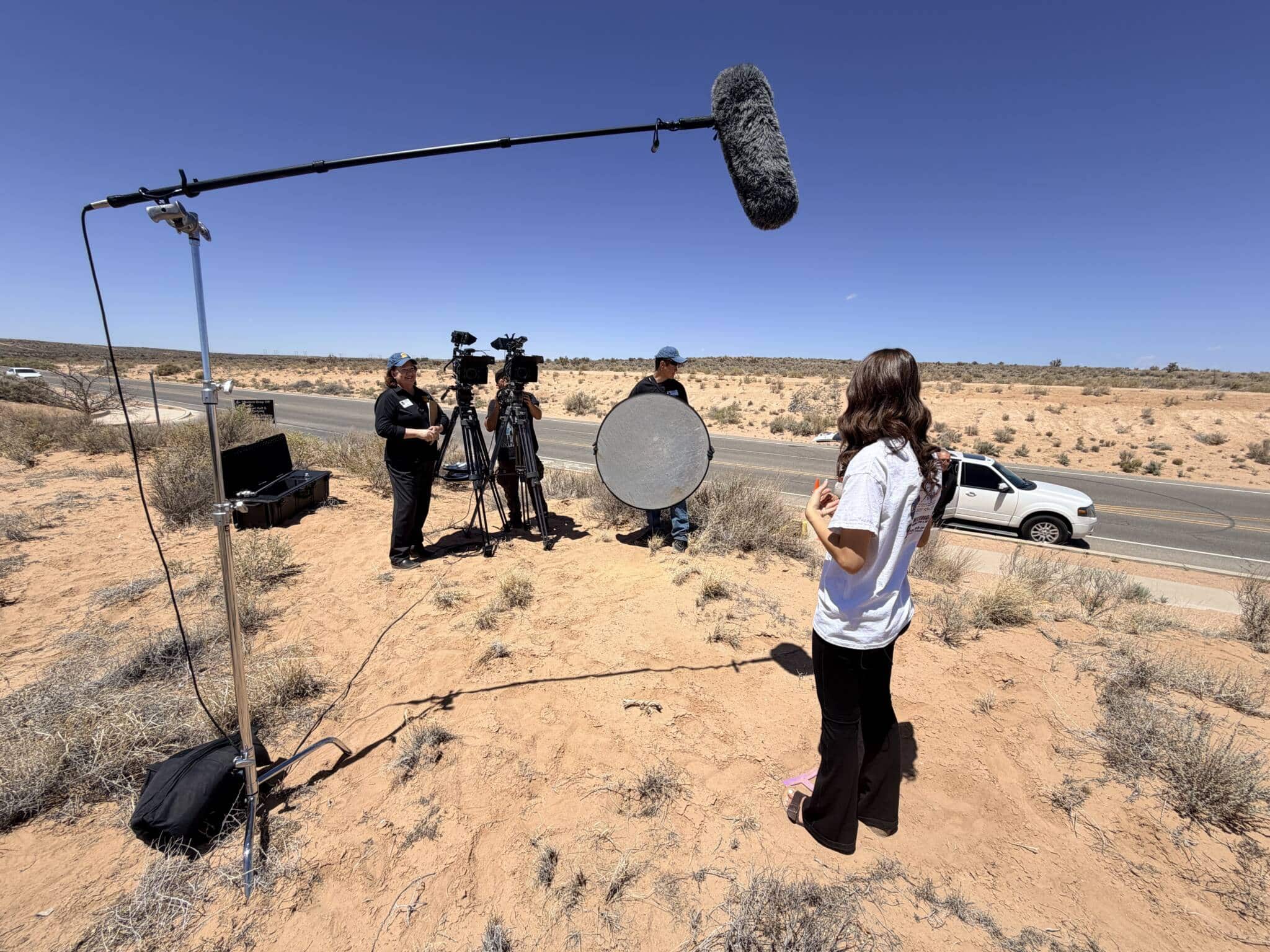 Film crew setting up outdoors in a desert: a long boom mic with furry windscreen over the scene, cameras on tripods, and people coordinating by a road.