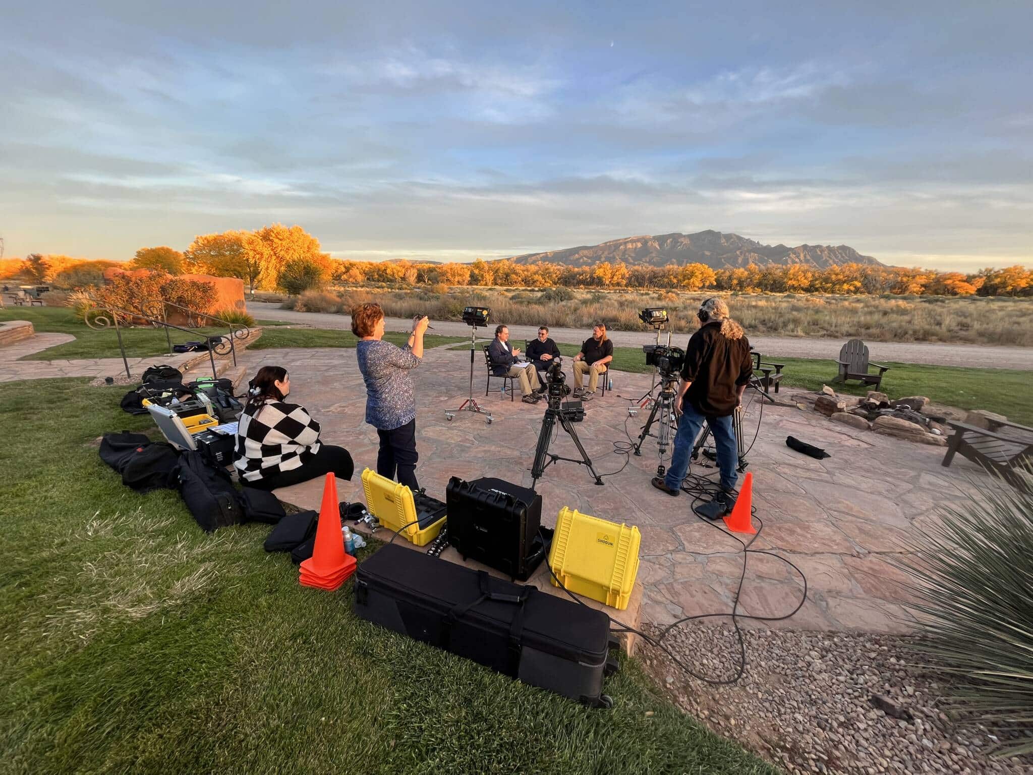 Crew filming outdoors at sunset with cameras, lighting gear, and protective cases on a paved patio; desert landscape and distant mountains in warm light.