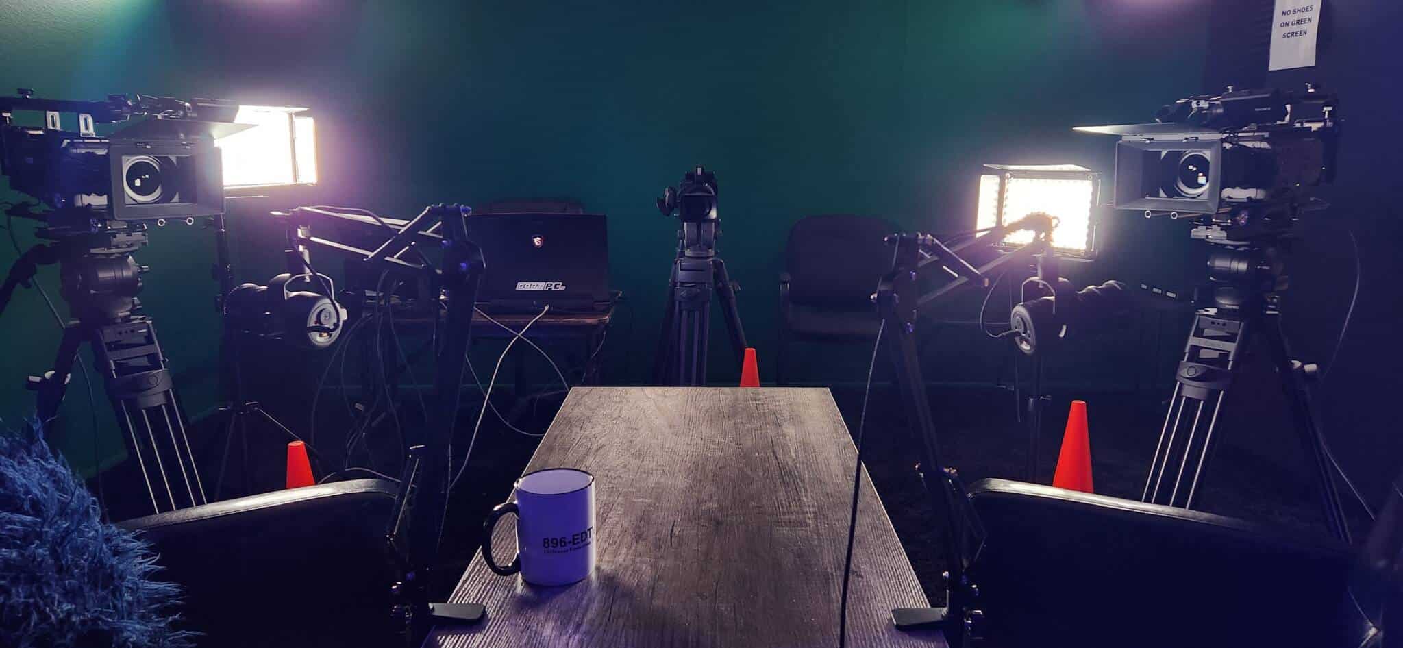 Video production studio with three lighting rigs on tripods facing a green screen; wooden table in foreground with a mug and cables scattered around.