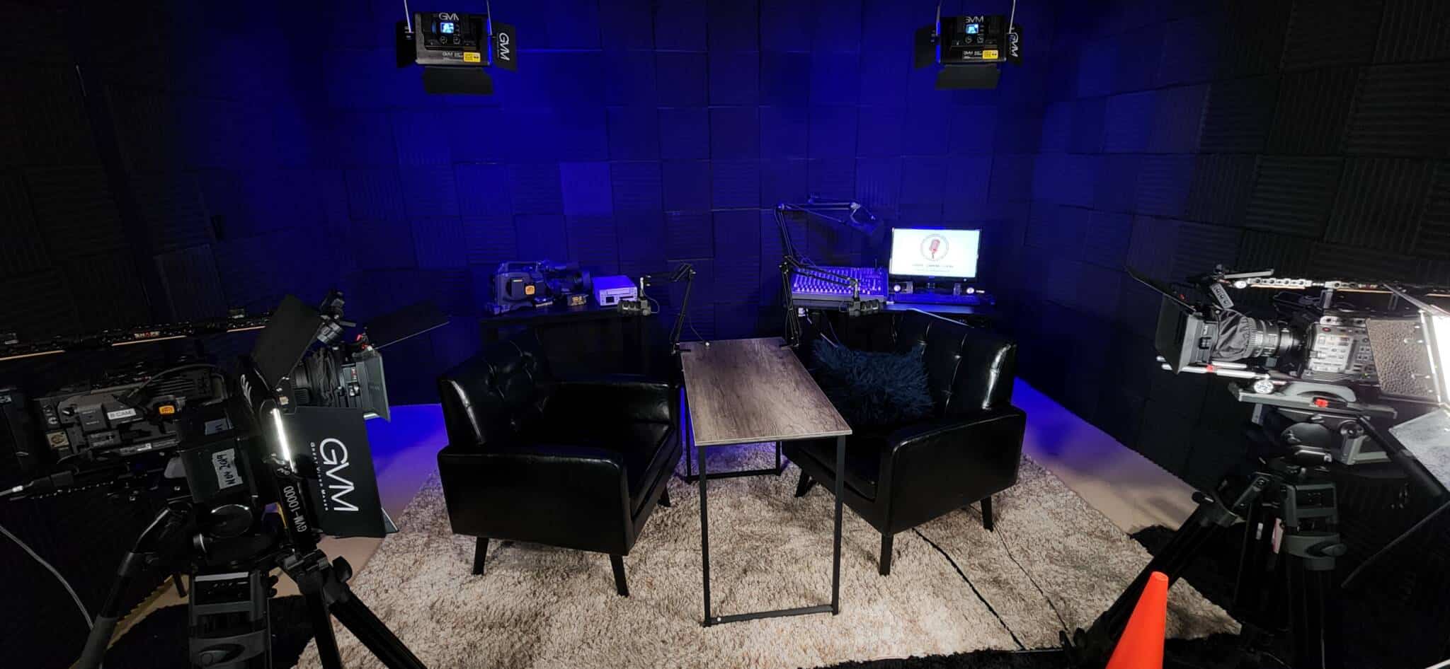 Blue acoustic-wall recording studio with a wooden table and four black chairs, surrounded by camera gear and a monitor for editing or streaming shows.