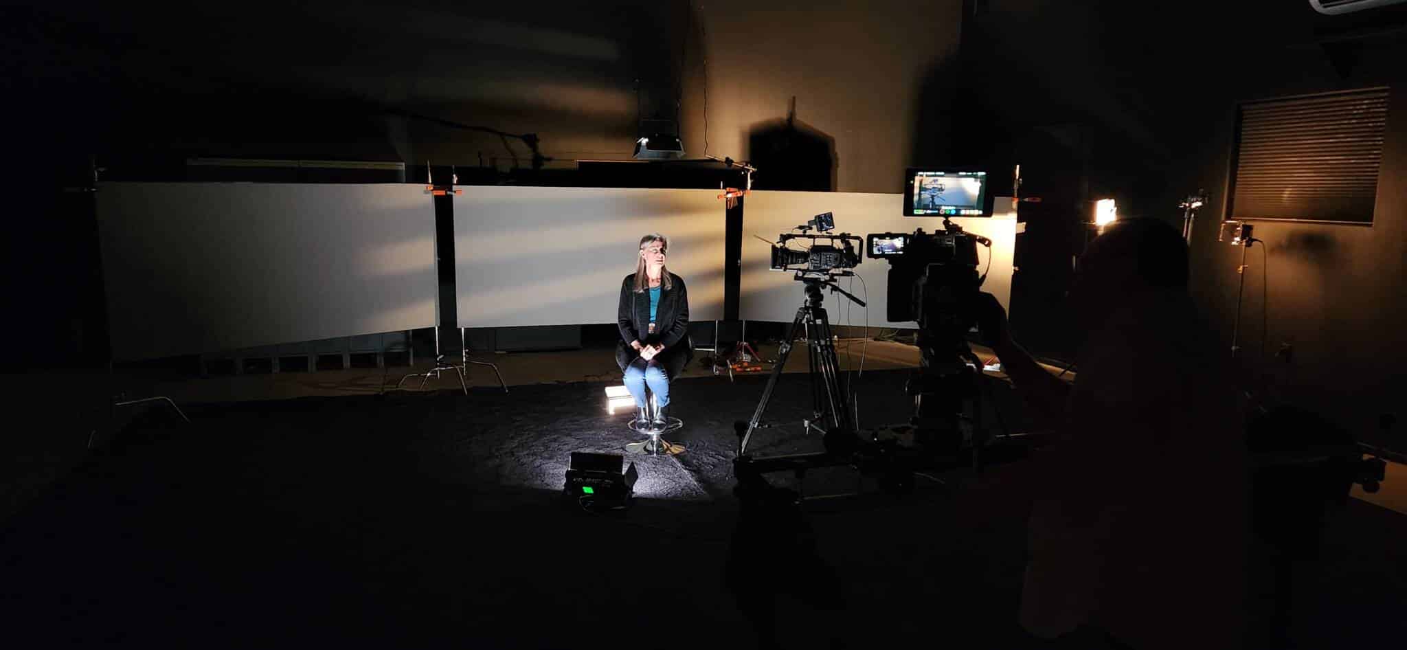 A woman sits on a stool in a dim video studio, surrounded by cameras, lights, and a backdrop behind her.