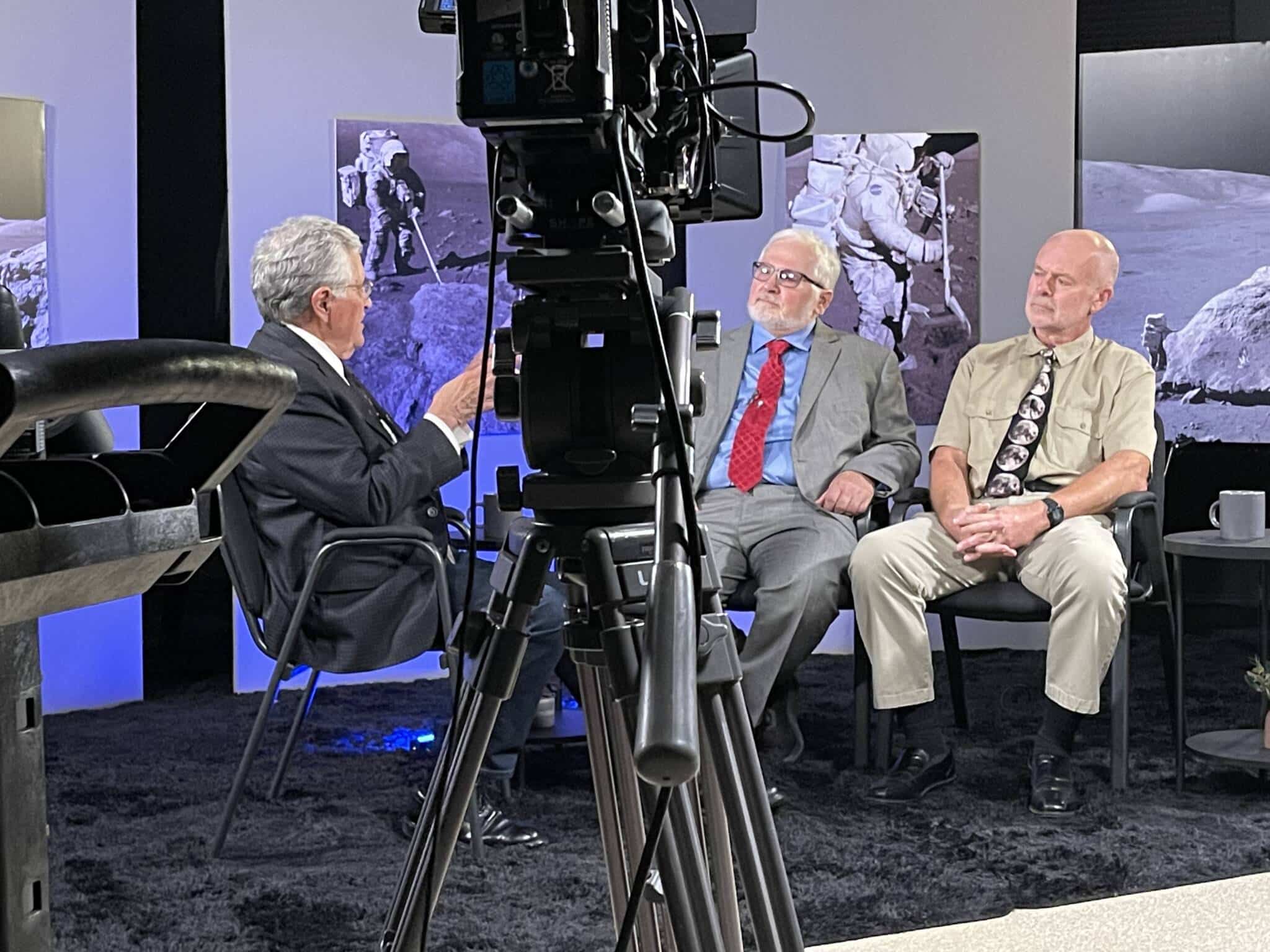 Three men in a studio discuss panel-style while a camera crane dominates the foreground; backdrop shows space-themed images of astronauts.
