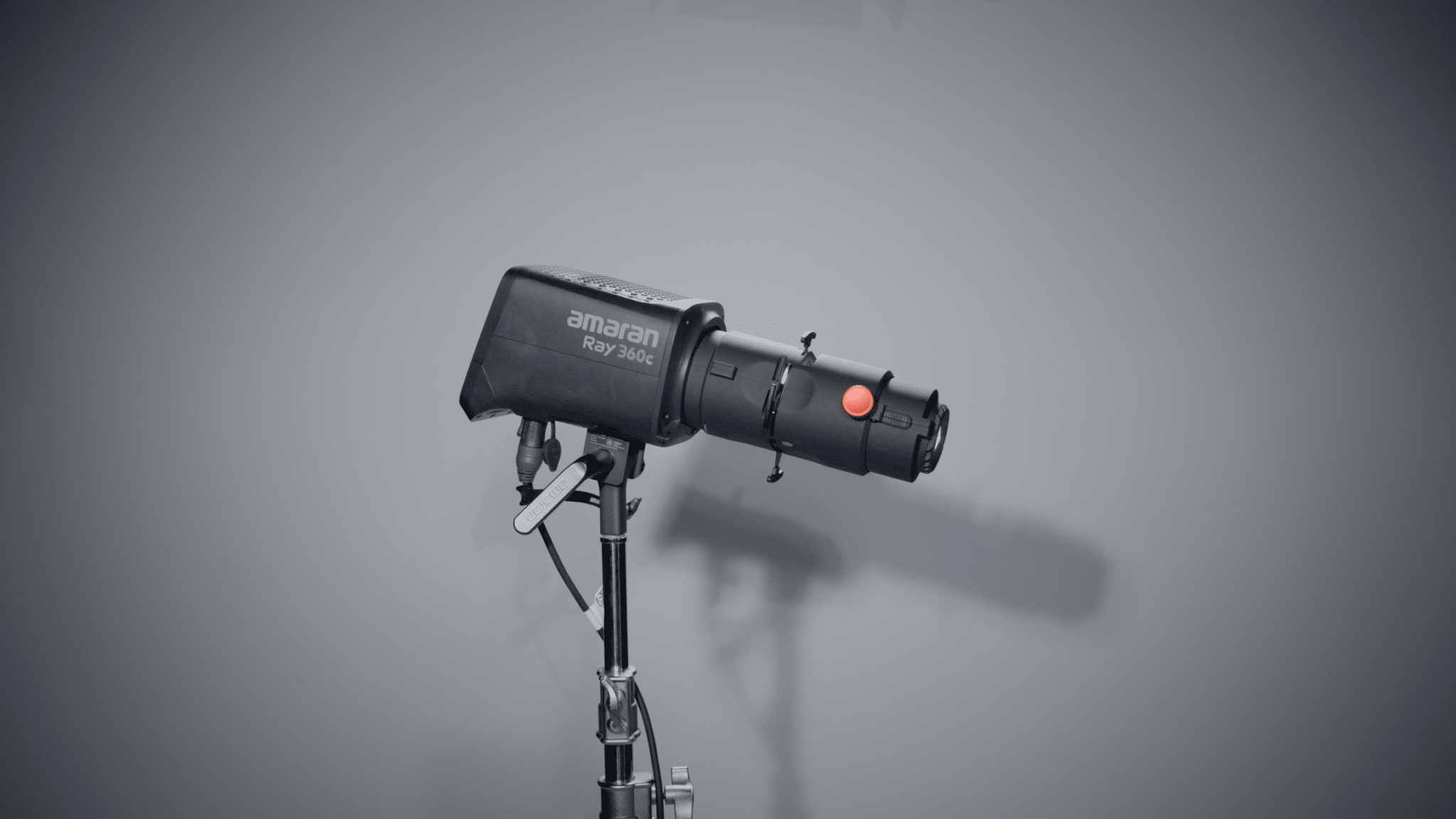 Studio strobe light on a stand facing left, with 'amaran Ray 360c' visible on the head in a neutral gray setup.