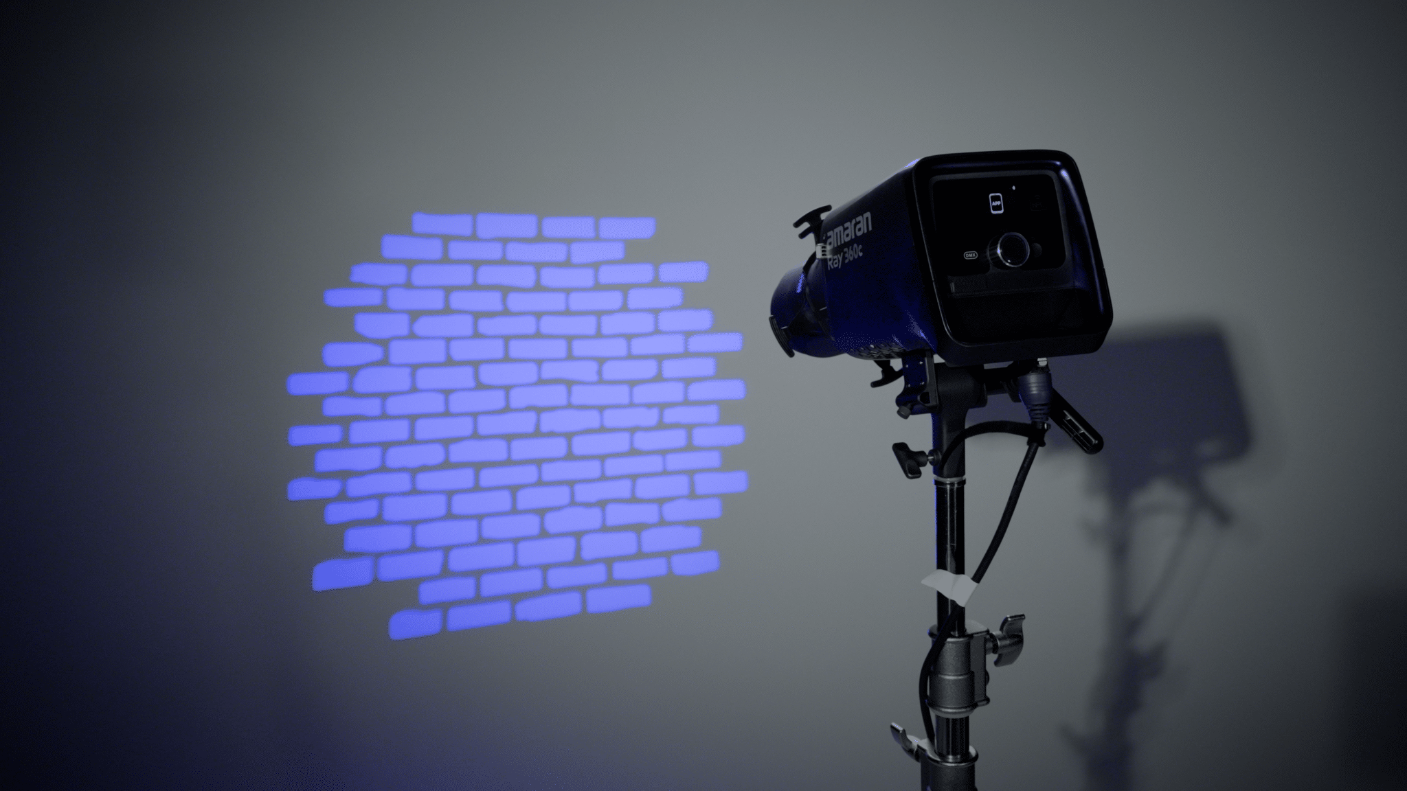 Studio lights on a stand cast a blue brick-pattern projection on a gray wall in a dim studio showpiece nearby.