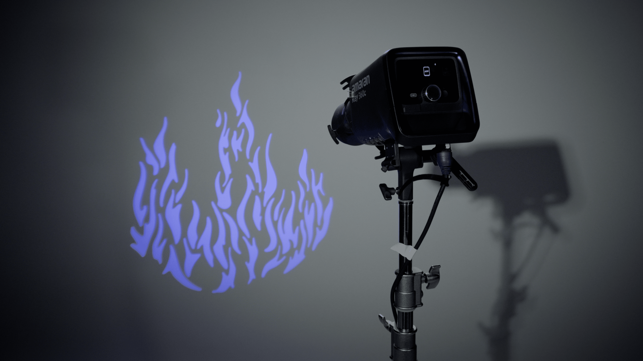 Studio light on a tripod projecting blue flame-shaped silhouettes onto a gray wall in a dim setting.