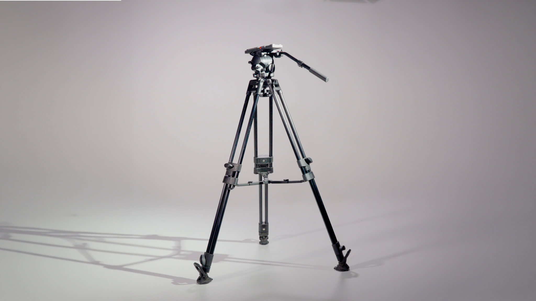 Professional studio tripod with a mounted camera head on a gray backdrop, legs spread and stabilizing on the floor.