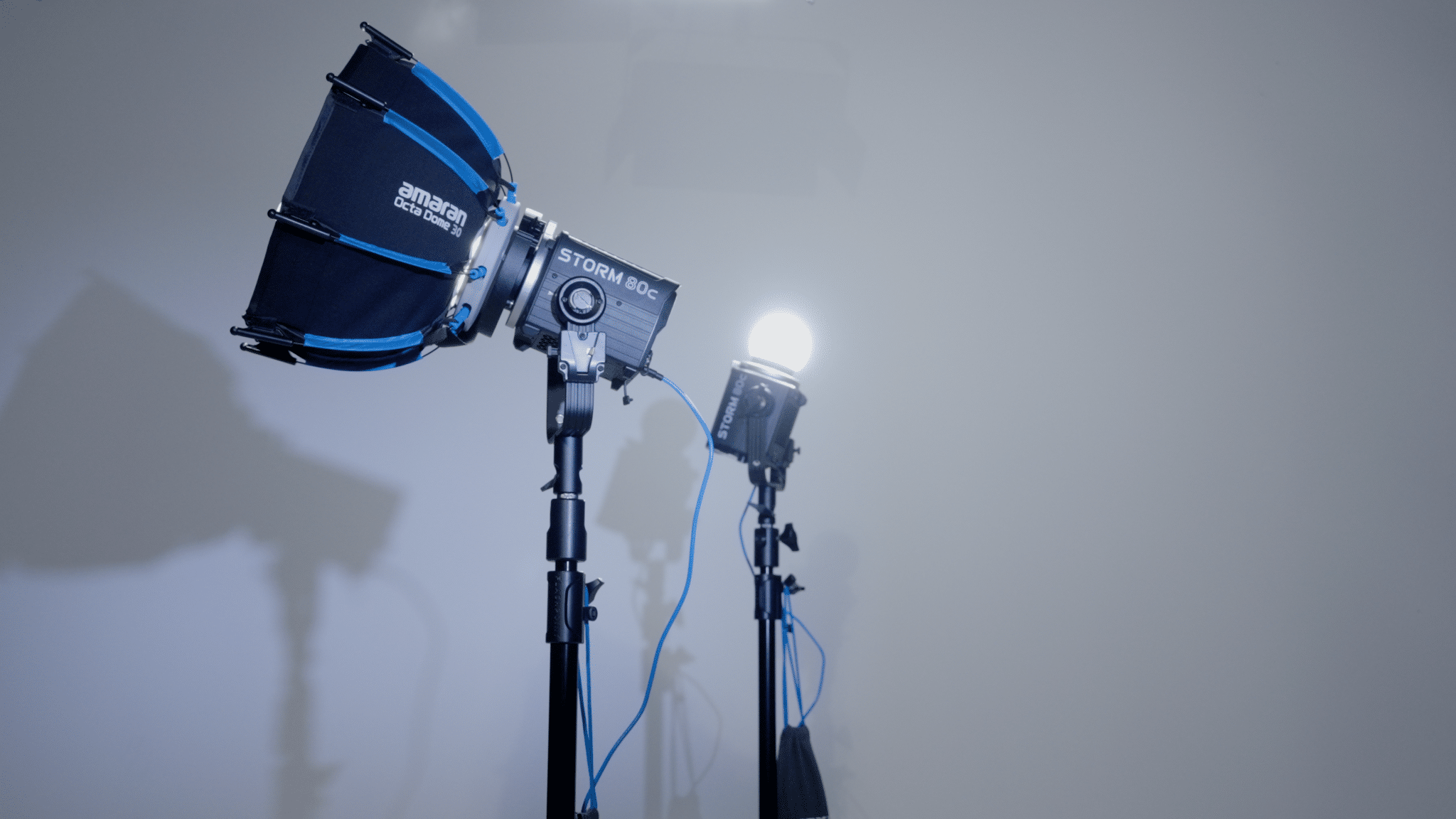 Close-up of two professional studio lights on stands with blue cables, one with a blue-accent softbox labeled Amaran, in a photo studio setup.