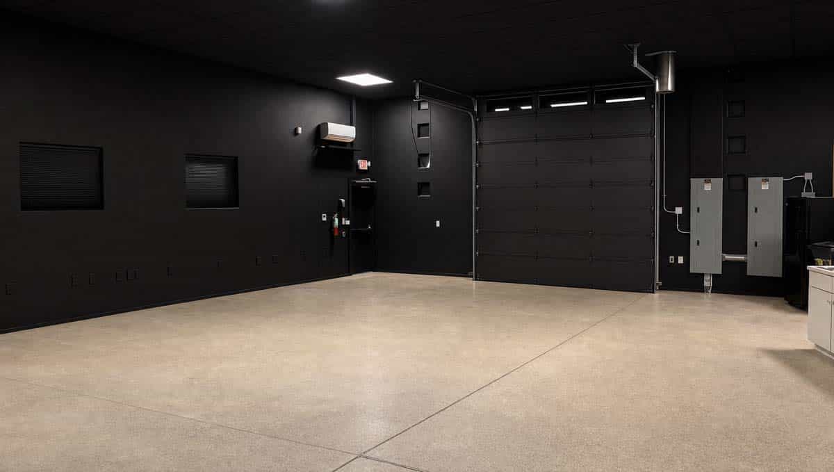 Empty black-walled industrial room with a large metal roll-up door and exposed electrical panels on the right.