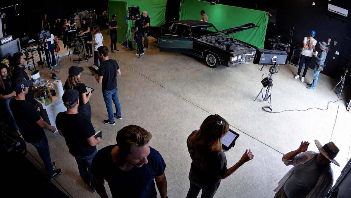 Film crew on a studio set around a black vintage car with its hood open, green screen backdrop and video gear nearby.