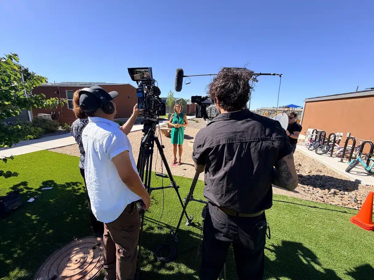 A woman in a green dress speaks on a sunny outdoor film set, surrounded by camera crew and equipment.