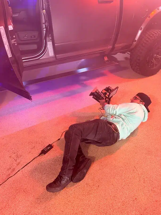 Person lying on their side on a garage floor, filming a pickup truck with a camera rig in a red and blue-lit scene.