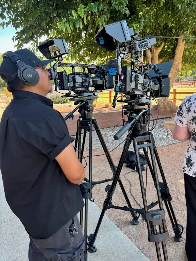 Two professional film cameras on tripods with a cameraman wearing headphones filming outdoors under trees.