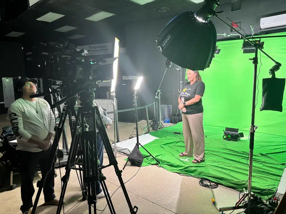 A woman stands on a green-screen stage in a studio, being filmed by a camera operator with a large camera and bright lights around.