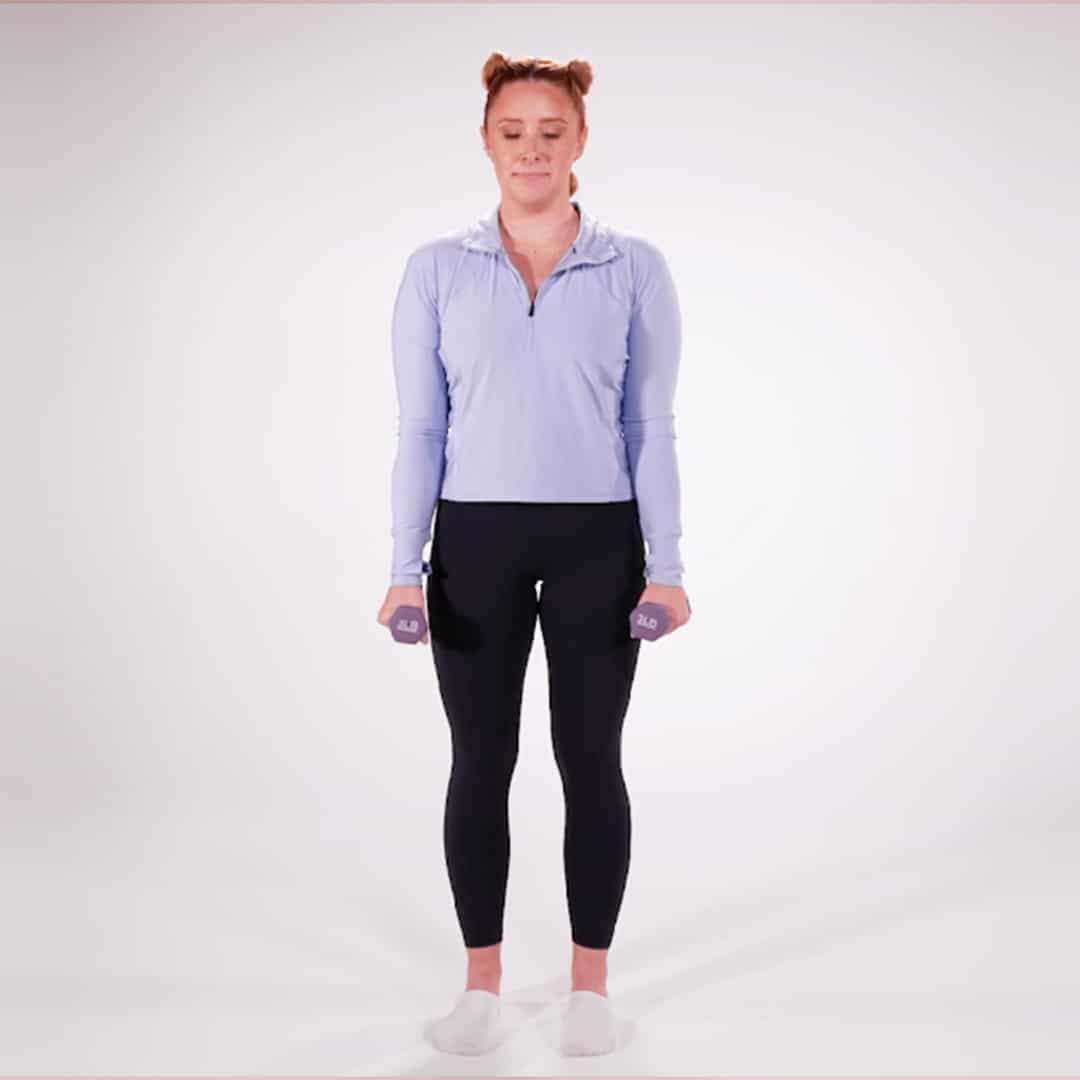Woman in a light blue long-sleeve athletic top and black leggings stands in a white studio, holding small purple dumbbells at her sides, ready to exercise.