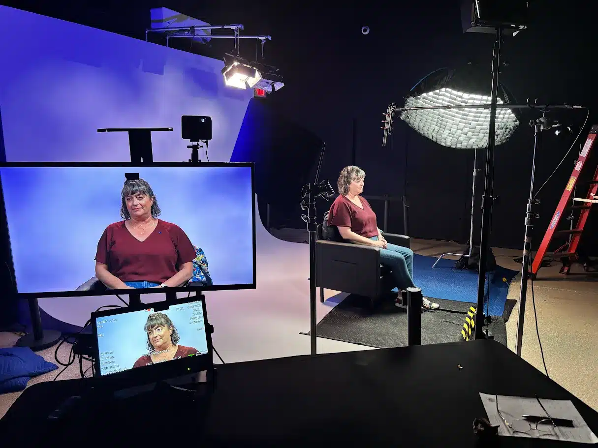 Woman in a maroon top sits in a black chair on a TV studio set with a blue backdrop and overhead lights visible behind her.